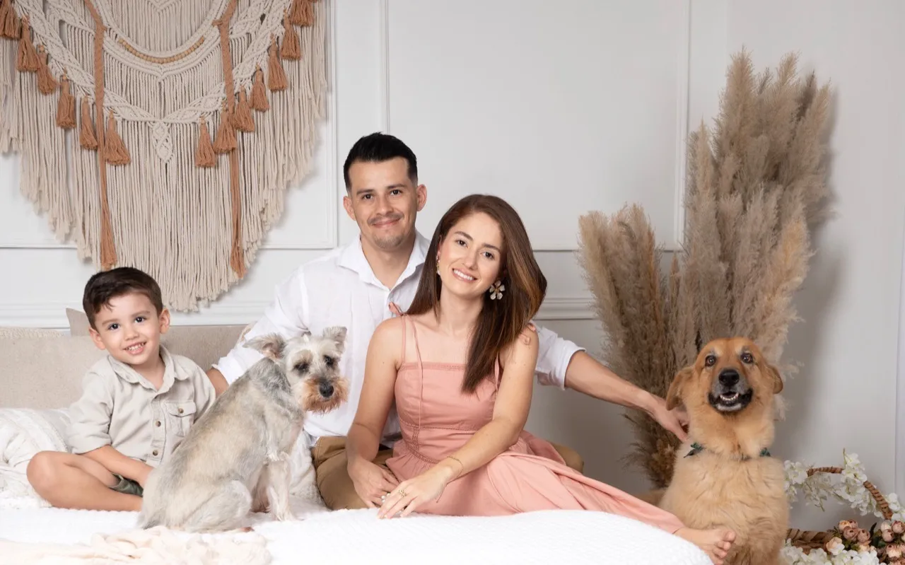 As duas pessoas que fazem a Nayra, sentadas em uma cama com o filho e os dois cachorros durante um ensaio de fotos em família.